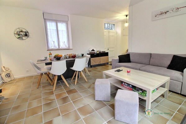 Un salon moderne avec un canapé gris, une table basse blanche et un coin repas avec des chaises blanches, et une cuisine en arrière-plan.
