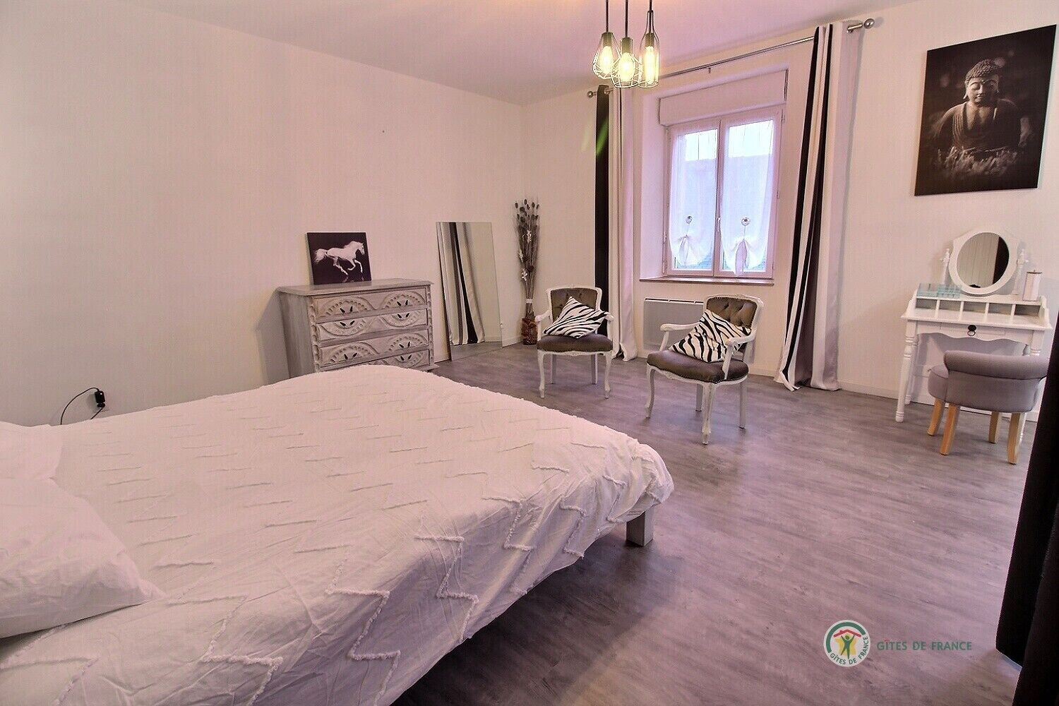 A bedroom with a bed, a dresser, a vanity with a mirror, two chairs, and a window with curtains.