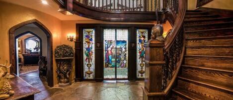 Una gran escalera con detalles de madera decorativos y un candelabro, que conduce a una habitación con piso de piedra y puertas de vidrio emplomado.