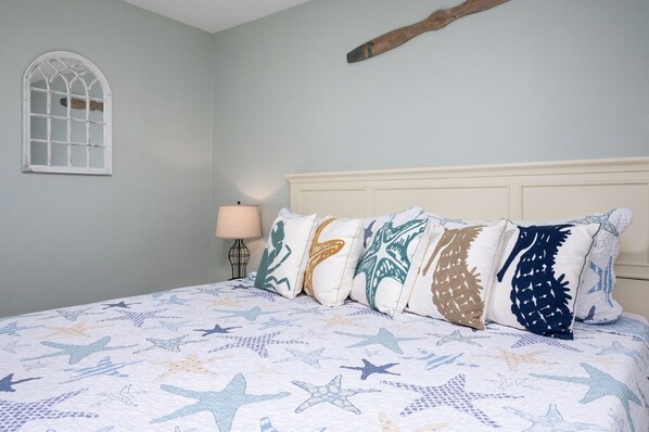 A bedroom with a bed featuring a starfish pattern bedspread, a wooden headboard, and a window with a white frame. - A Lil' Tipsea 4 Bedroom Home by RedAwning (Port Bolivar)
