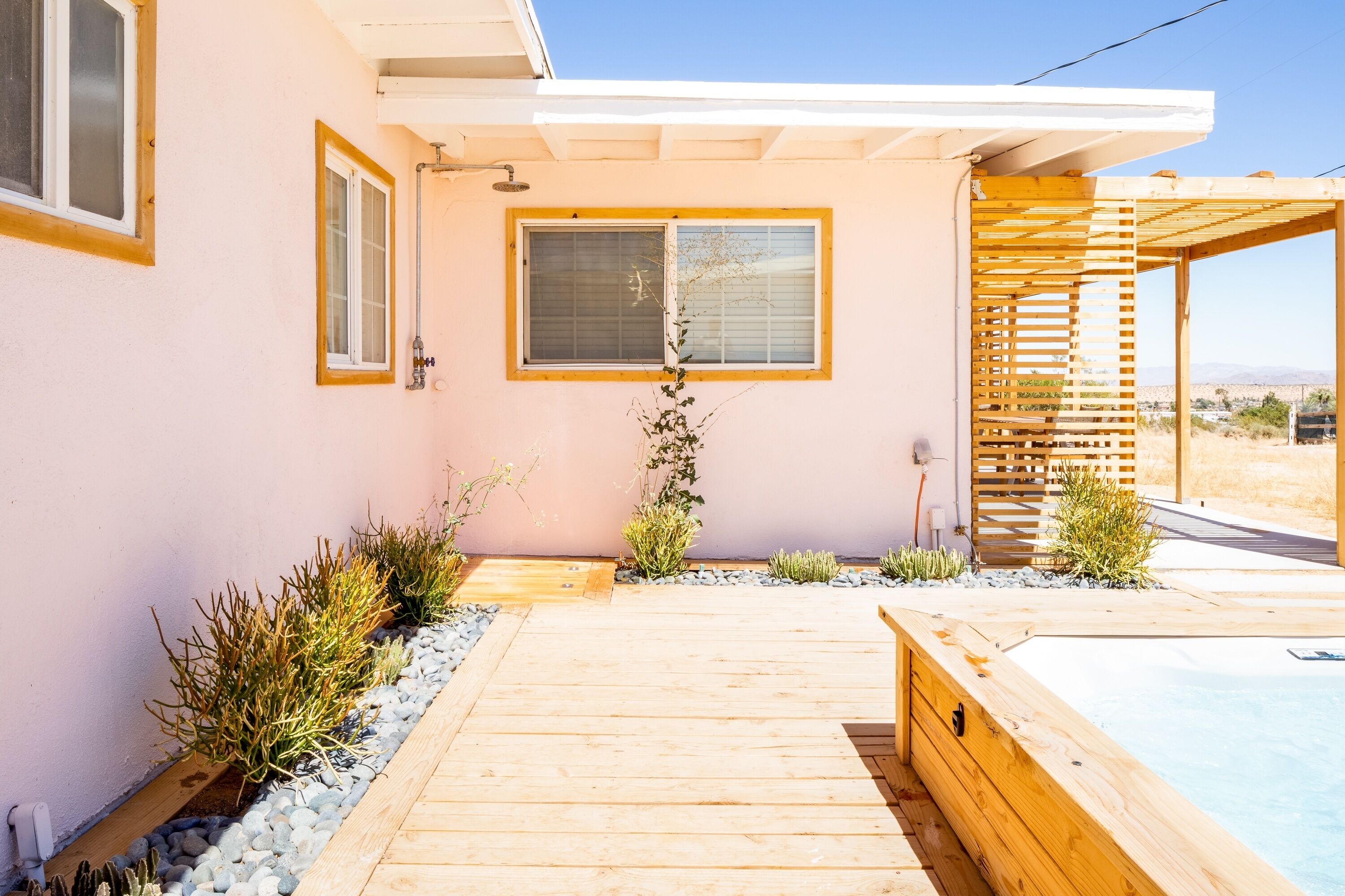 A modern house with a wooden deck leading to a pool, surrounded by plants and a pergola.