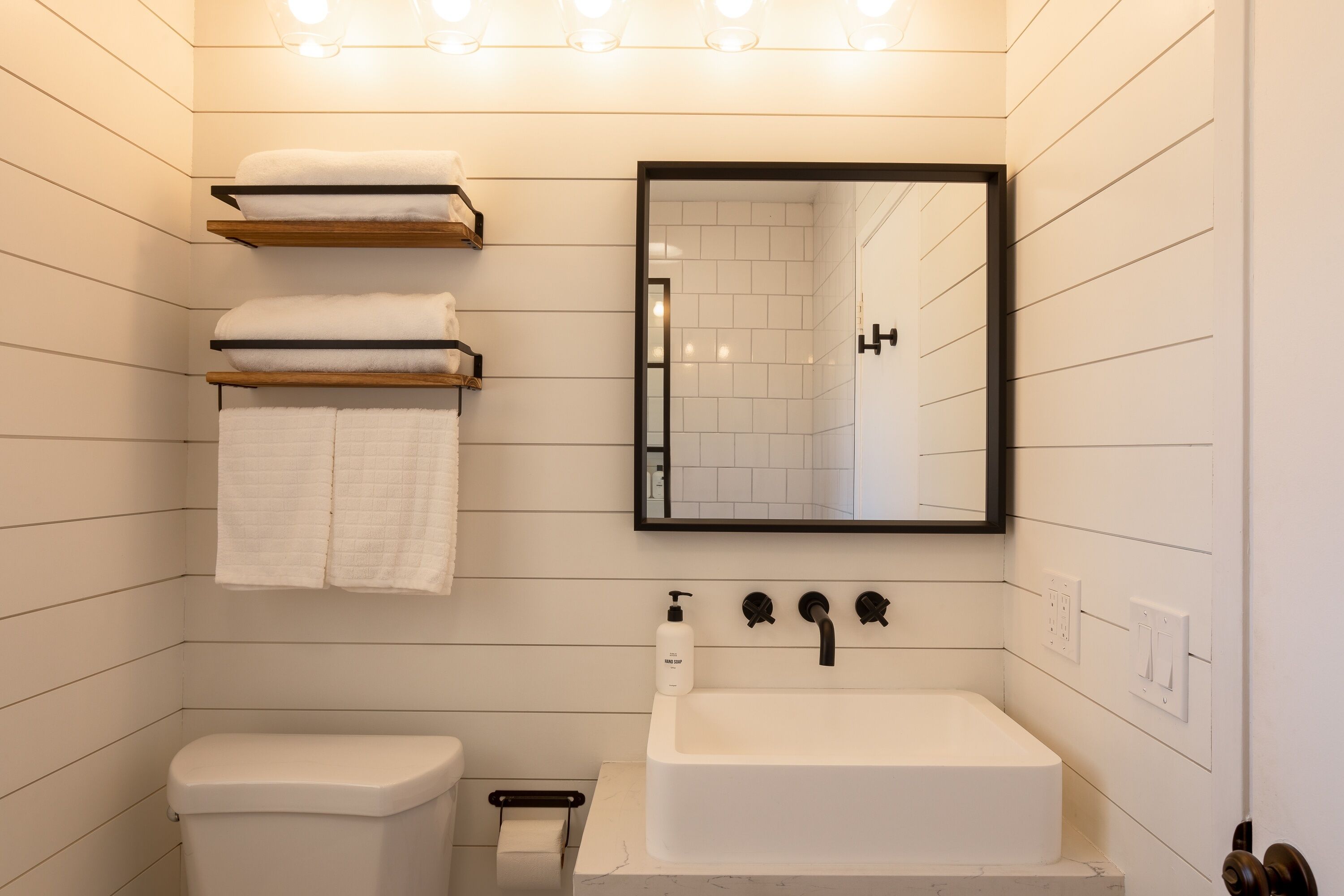 Une salle de bain moderne avec un lavabo blanc, un miroir et des toilettes. On trouve des serviettes sur une étagère et un distributeur de savon sur le lavabo.