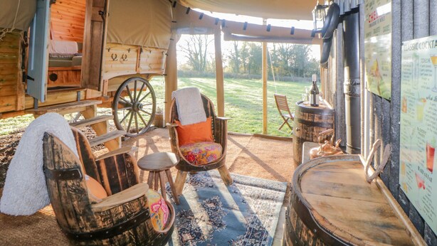 A rustic tented room with a wooden bed, a large wheel, a chair with a colorful cushion, and a bar area with a menu.
