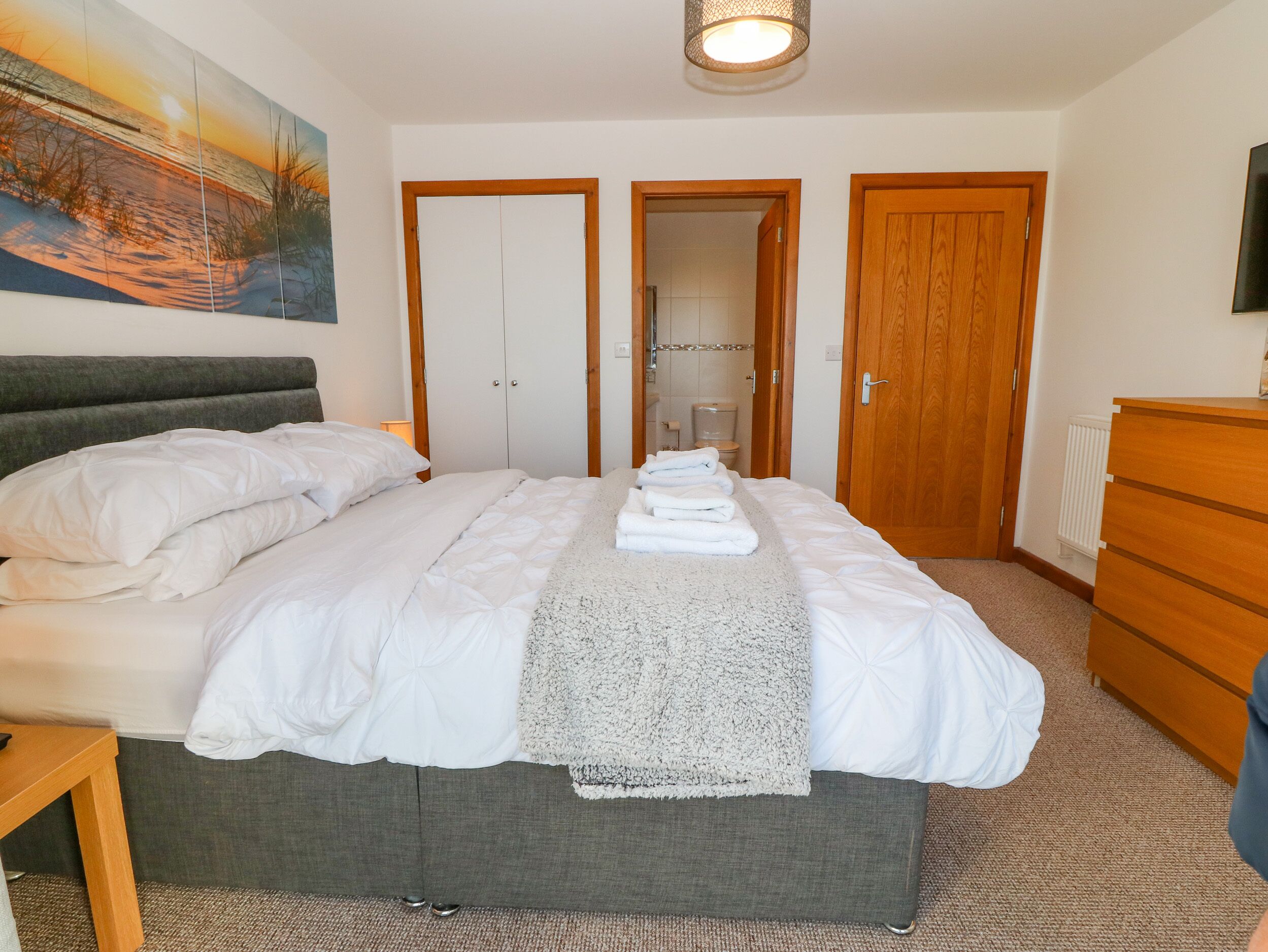 A neatly made bed with white linens and a grey headboard, a wooden dresser, and a painting of a sunset on the wall.