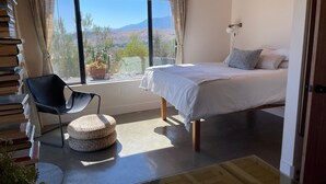 Quarto com cama, cadeira, mesinha e janela com vista para paisagem desértica.