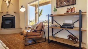 A living room with a fireplace, a leather chair, a wooden shelf with books and a typewriter, and a large clock on the wall. - Horizon House Hot Tub, Fire Pit & Bbq Adobe In Jt 4 Bedroom Home by RedAwning (Joshua Tree)