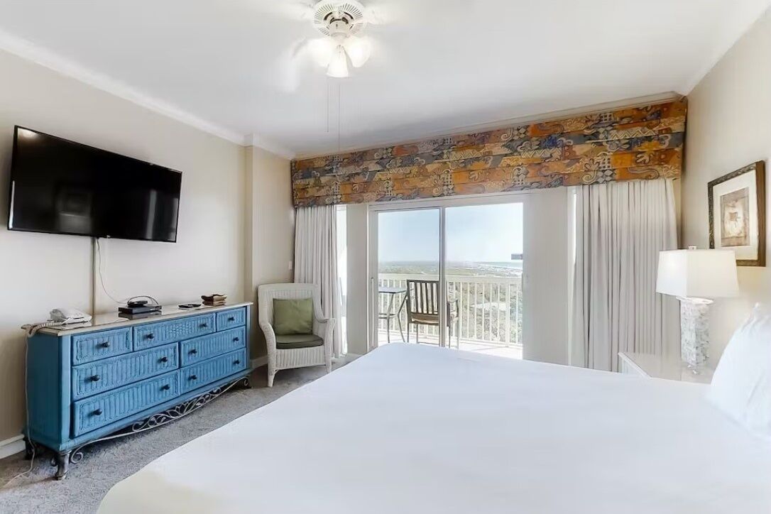 Ein Hotelzimmer mit einem großen Bett, einer blauen Kommode, einem Flachbildfernseher, einem Sessel, einem Balkon mit Blick und einem Deckenventilator.