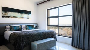A modern bedroom with a large bed, a bench, and a view of a building through a large window. - The Capital 20 West (Johannesburg)
