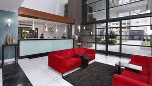 A modern hotel lobby with a reception desk, red sofas, and large windows offering views of the outside. - The Capital 20 West (Johannesburg)
