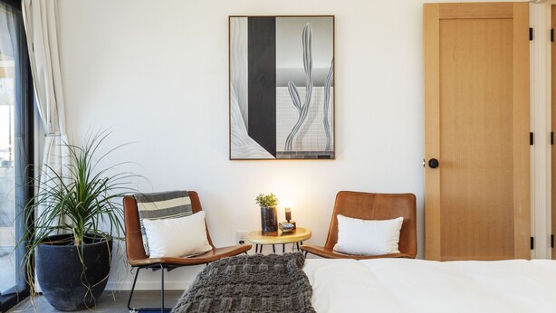 A hotel room with a bed, two chairs, a small table with a plant, a potted plant, and a painting on the wall. - Twilight Terrace - Relax In Style 2 Bedroom Home by RedAwning (Joshua Tree)