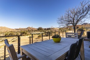 An outdoor patio with a wooden table and chairs, a yellow planter, and a view of a desert landscape with mountains in the background. - Twilight Terrace - Relax In Style 2 Bedroom Home by RedAwning (Joshua Tree)