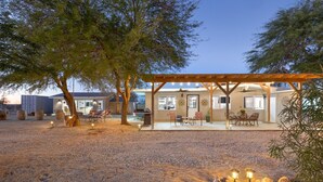 A covered outdoor patio with seating and a dining area, surrounded by trees and a gravel ground. - Mystic Views - Pool, Hot Tub, Game Room, Fire Pit & Desert Views 4 Bedroom Home by RedAwning (Twentynine Palms)