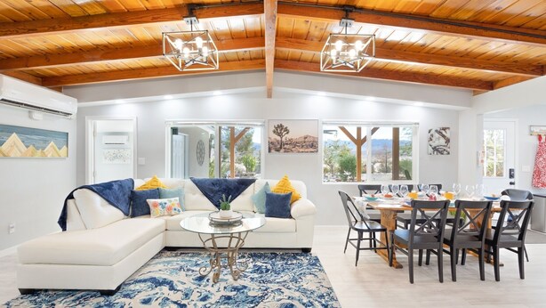 A spacious living area with a white sofa, a round coffee table, and a dining table set for six. The room features wooden ceiling beams, large windows with a view, and a blue patterned rug. - Mystic Views - Pool, Hot Tub, Game Room, Fire Pit & Desert Views 4 Bedroom Home by RedAwning (Twentynine Palms)