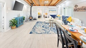 A modern living room with a fireplace, a sofa, a dining table set, and a large window. - Mystic Views - Pool, Hot Tub, Game Room, Fire Pit & Desert Views 4 Bedroom Home by RedAwning (Twentynine Palms)
