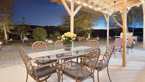 An outdoor patio with a round glass table and wicker chairs, surrounded by a wooden pergola with string lights. - Mystic Views - Pool, Hot Tub, Game Room, Fire Pit & Desert Views 4 Bedroom Home by RedAwning (Twentynine Palms)