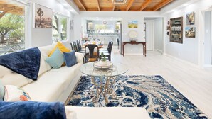 A spacious living room with a white sofa, a glass coffee table, and a blue patterned rug. There is a dining area with a wooden table and chairs, and a kitchen in the background. - Mystic Views - Pool, Hot Tub, Game Room, Fire Pit & Desert Views 4 Bedroom Home by RedAwning (Twentynine Palms)