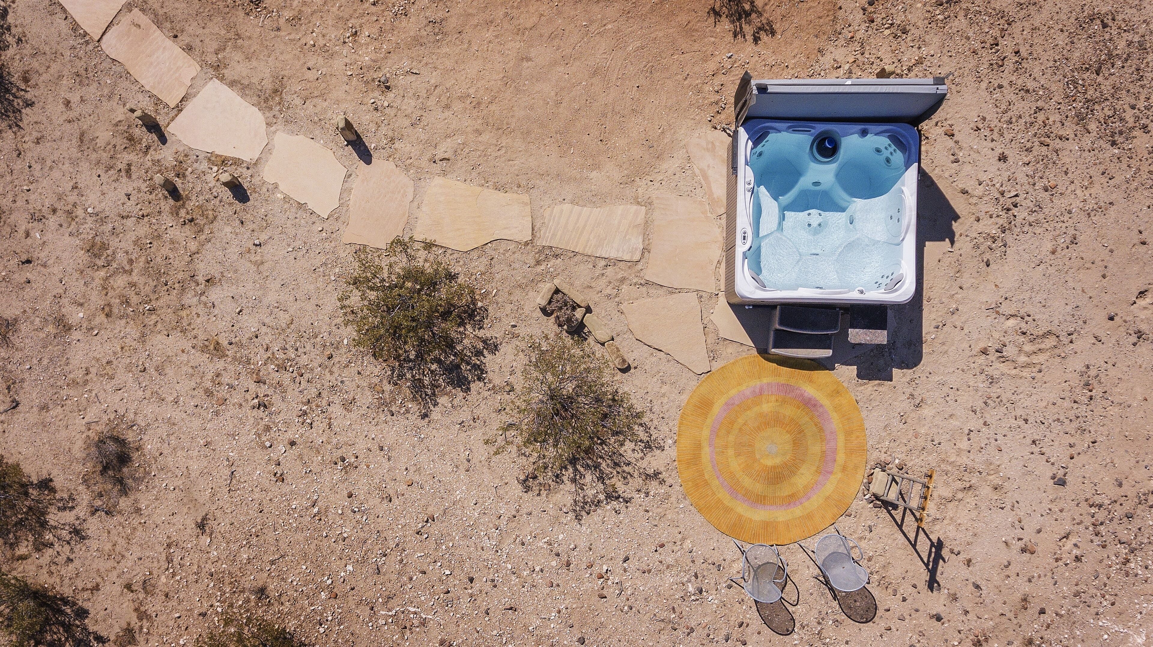 An aerial view of a hot tub with a circular seating area and a small table, surrounded by a gravel path and sparse vegetation.