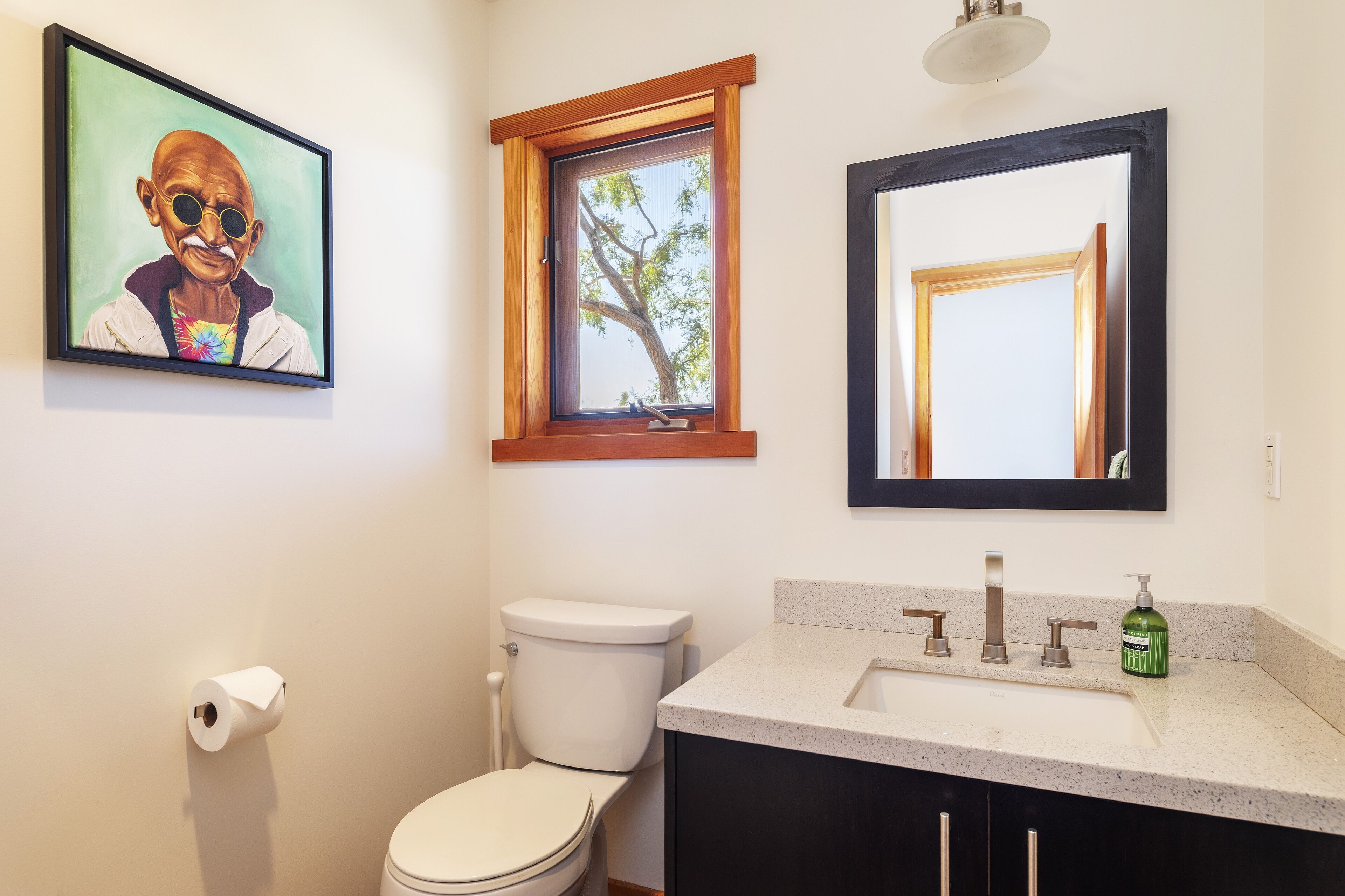 Ein Badezimmer mit Toilette, Waschbecken mit Spiegel, einem gerahmten Bild an der Wand und einem Fenster mit Blick auf Bäume.