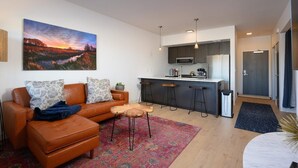 A modern living room with a brown leather sofa, a coffee table, and a colorful rug. A kitchen with dark cabinets and stainless steel appliances is visible in the background. - Stylish SOBO Downtown Condo - Steps to Main St. (Bozeman)