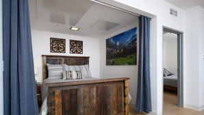 A bedroom with a wooden bed, a night sky painting, and a view into another room. - Stylish SOBO Downtown Condo - Steps to Main St. (Bozeman)