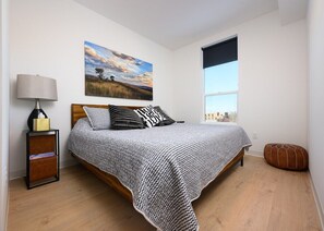 A bedroom with a bed, a nightstand, a lamp, a window, and a painting on the wall.