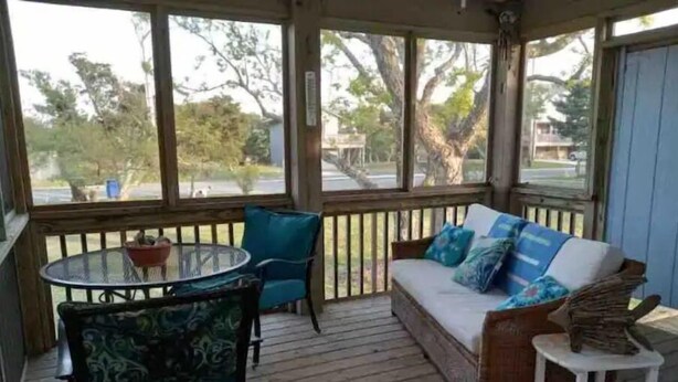 A covered porch with a wooden deck, a round glass table, two chairs, a sofa with cushions, and a small white stool. - Eagle Pass Cabina- Unique Cabin Perfect for Two! (Hatteras)