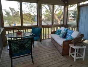 A covered porch with a wooden deck, a round glass table, two chairs, a sofa with cushions, and a small white stool.