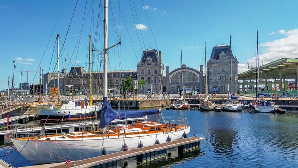 Un port esportiu ple de vaixells amarrats, un edifici gran amb cúpula i un pont al fons.