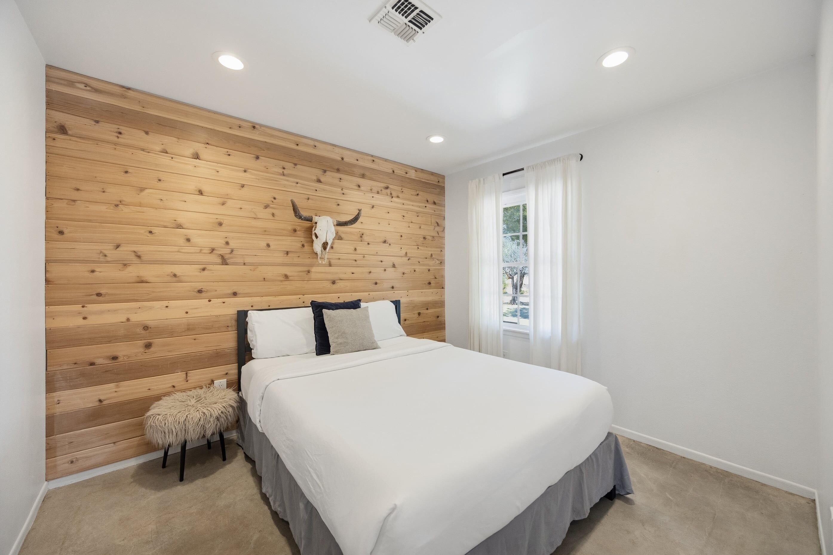 A bedroom with a wooden headboard, a bed with white bedding, a small stool, and a window with curtains.