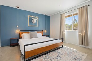 A bedroom with a bed, bedside tables, a hanging light fixture, a framed skull decoration, and a large window with curtains. - Cosmic Rabbit - Family Fun With Hot Tub!! 3 Bedroom Home by RedAwning (Joshua Tree)