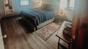 A bedroom with a wooden ceiling, a bed with a blue and white quilt, a nightstand with a lamp, and a window with blinds. - Sand Life - Pelican's Southern Nest! - Pet Friendly! (Kill Devil Hills)