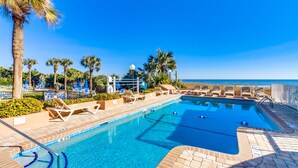 A clear blue pool with lounge chairs and palm trees, surrounded by a fence and a view of the ocean.