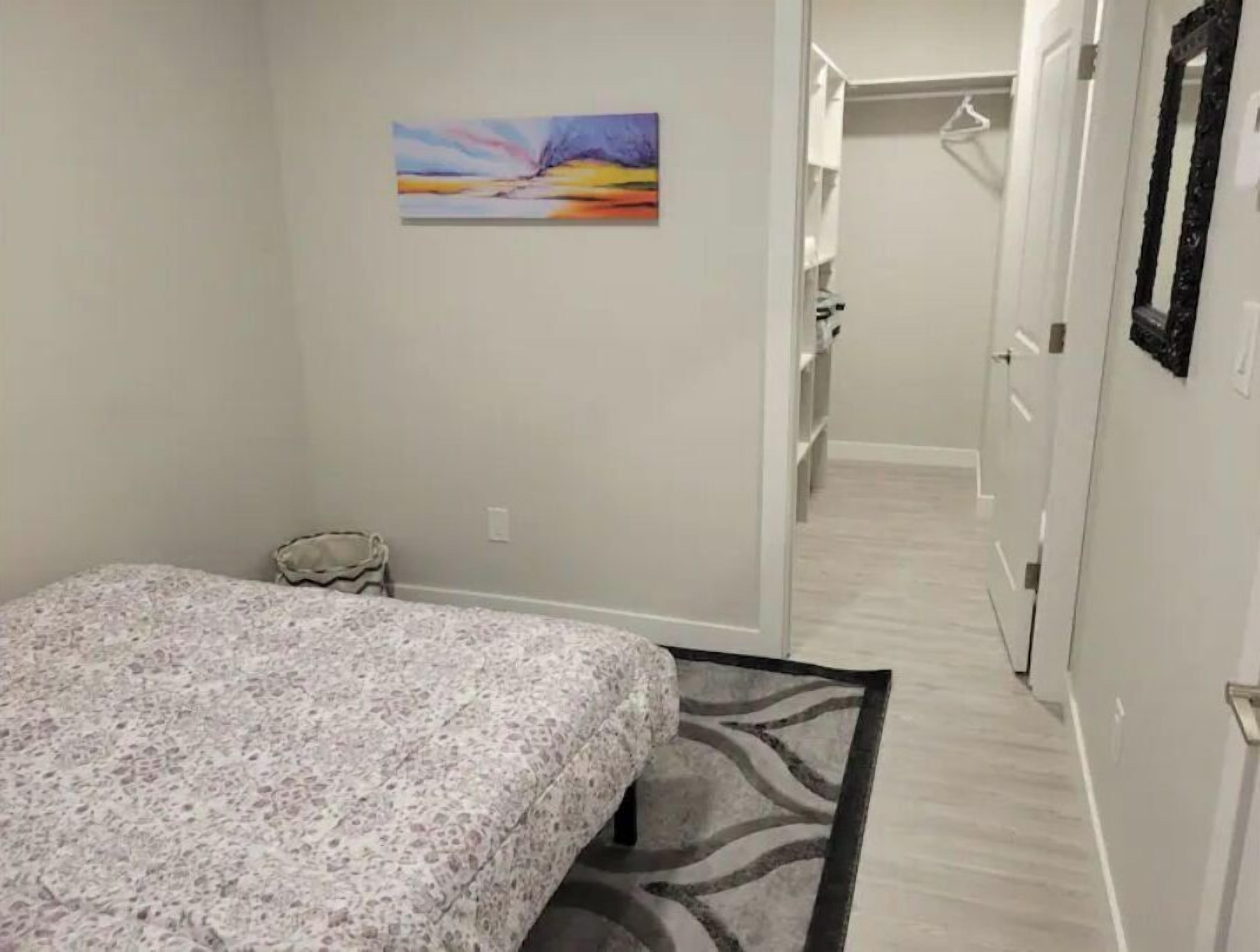 Une chambre avec un lit, une petite table de chevet, une chaise, un tableau accroché au mur et un couloir menant à une salle de bain.
