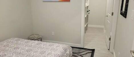 Une chambre avec un lit, une petite table de chevet, une chaise, un tableau accroché au mur et un couloir menant à une salle de bain.