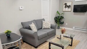 A living room with a grey sofa, a glass coffee table, a small round table with chairs, a potted plant, and a flat-screen TV mounted on the wall. - Grande Royale (Saskatoon)