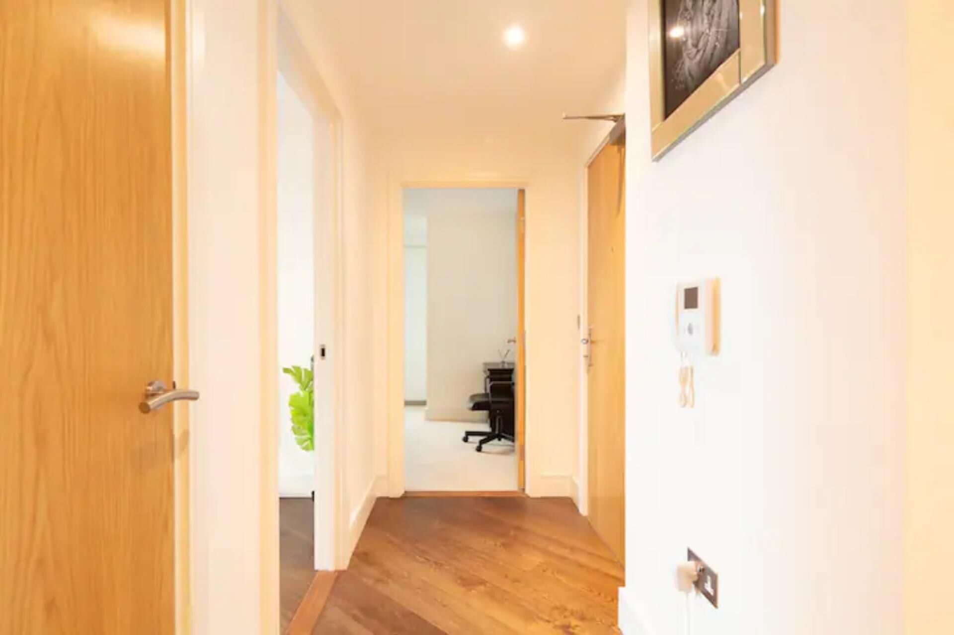 A hallway with wooden flooring, a door on the left, and a wall-mounted light fixture on the right.
