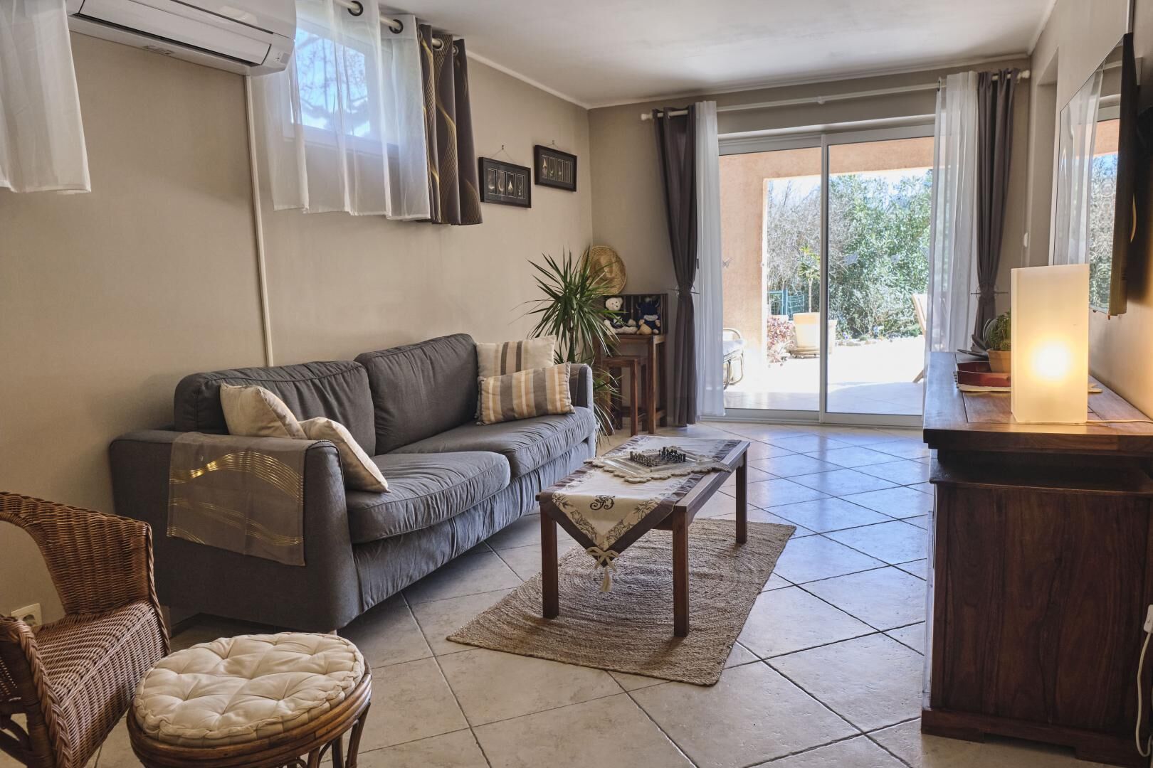 A living room with a grey sofa, a wooden coffee table, a wicker chair, and a cushioned ottoman. There is a large window with curtains, a plant, and a lamp on a wooden side table.
