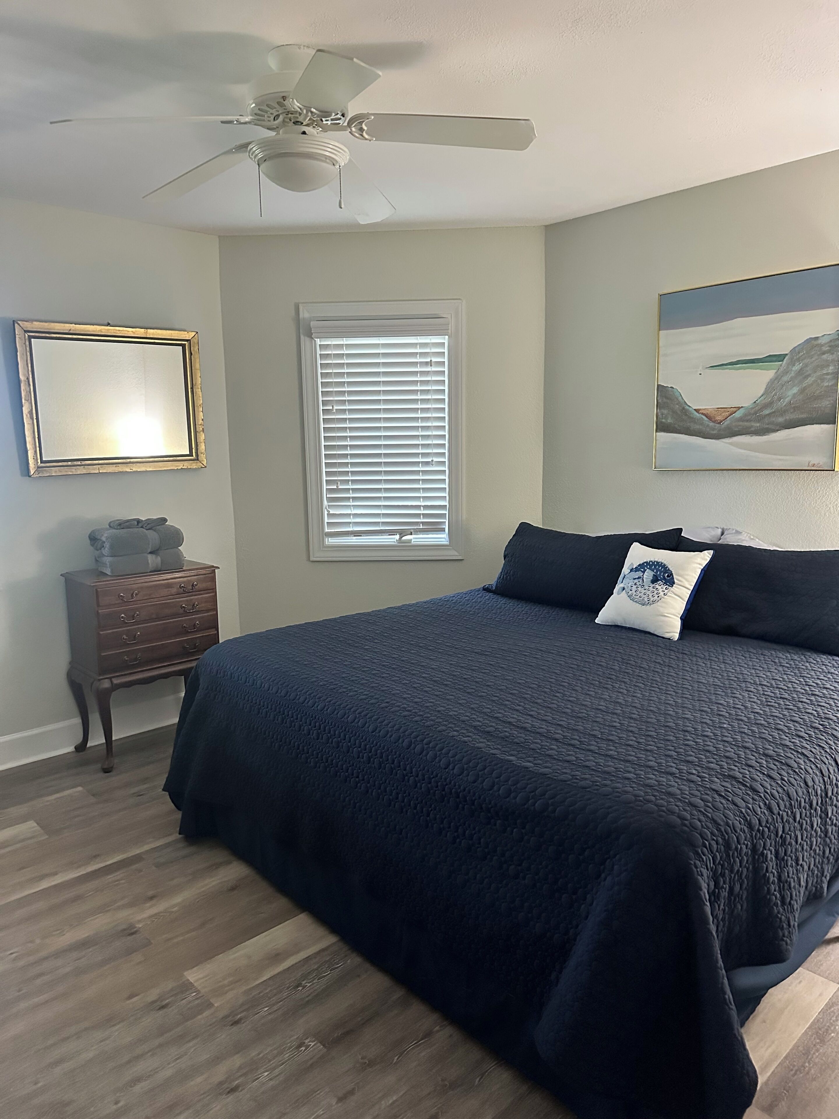 A bedroom with a bed, a ceiling fan, a dresser, a window with blinds, and a painting on the wall.