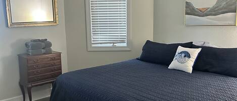 A bedroom with a bed, a ceiling fan, a dresser, a window with blinds, and a painting on the wall.