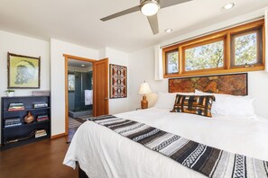 A bedroom with a large bed, a ceiling fan, a window with a view of trees, a bookshelf, and a door leading to a bathroom. - Constellation Compound - Your Own Private 10 Acres 4 Bedroom Home by RedAwning (Joshua Tree)