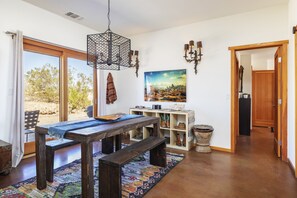 A spacious room with a wooden table and bench, a hanging light fixture, a bookshelf, and a large window with a view of the outdoors. - Constellation Compound - Your Own Private 10 Acres 4 Bedroom Home by RedAwning (Joshua Tree)