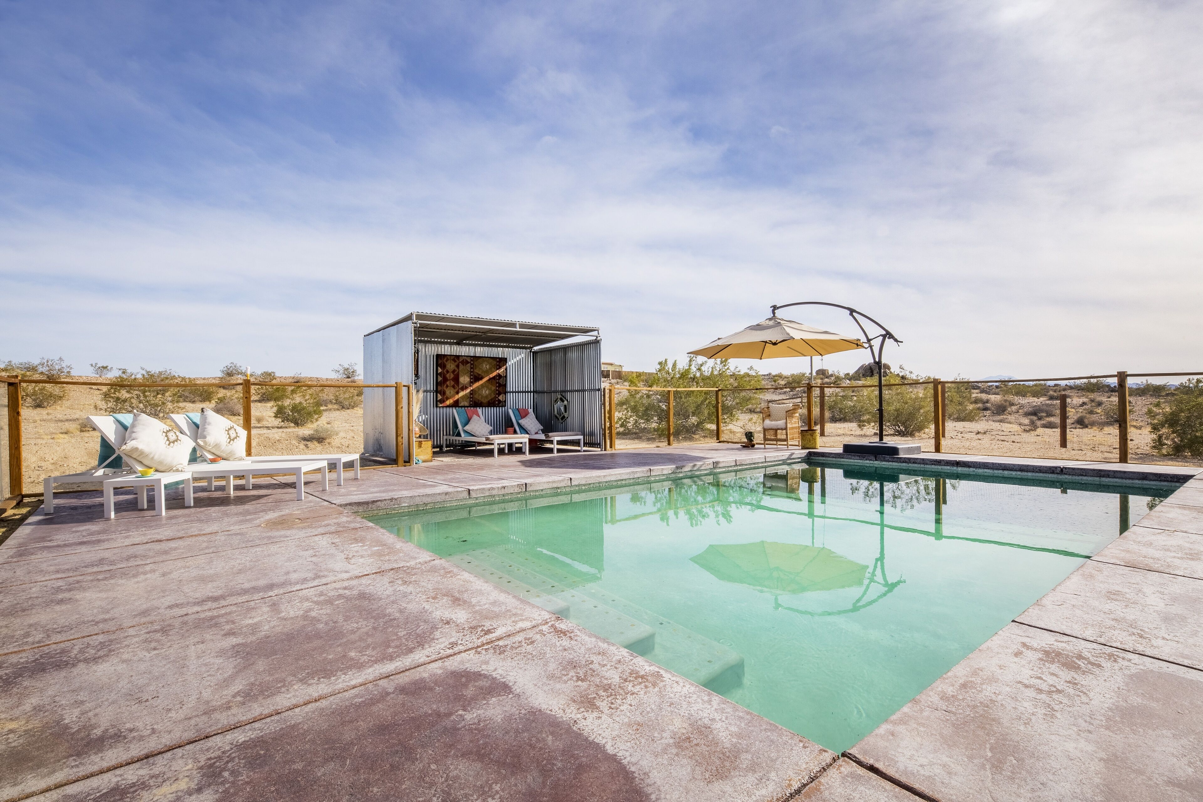 A pool area with lounge chairs and a small building, surrounded by a fence and desert landscape.