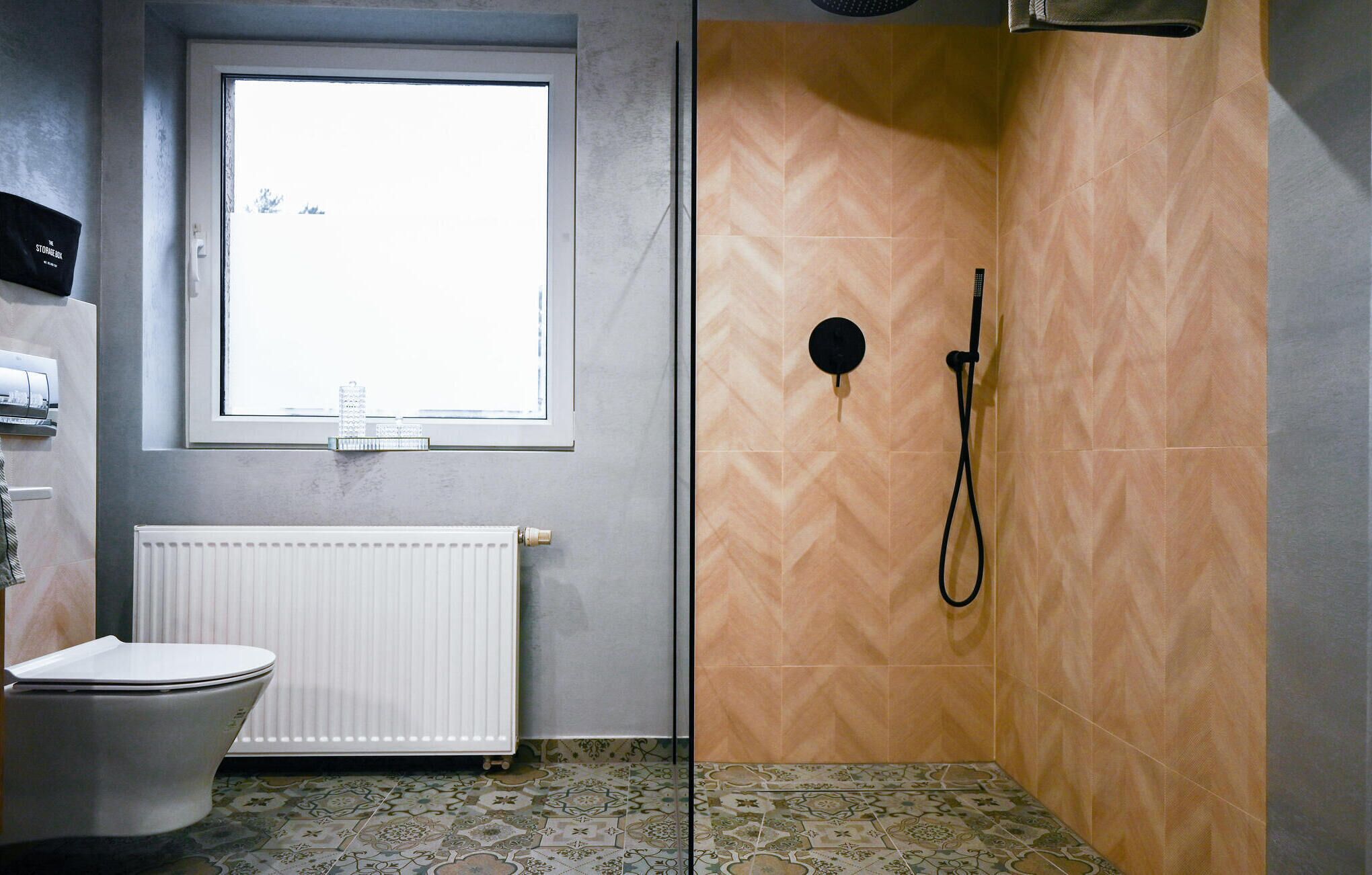 Un bagno con il water, una finestra, una doccia con tubo nero e piastrelle a motivi.