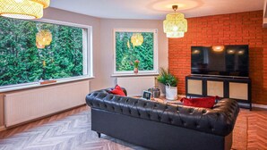 Uma sala de estar com um sofá de couro preto, duas janelas com vista para o verde, uma parede de tijolos, uma televisão e luminárias de vime.