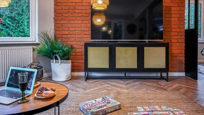 Uma sala de estar aconchegante com parede de tijolos, uma TV de tela plana presa acima de um móvel preto com painéis perfurados, um laptop, um copo de vinho, um jogo de Monopoly e um vaso de planta.