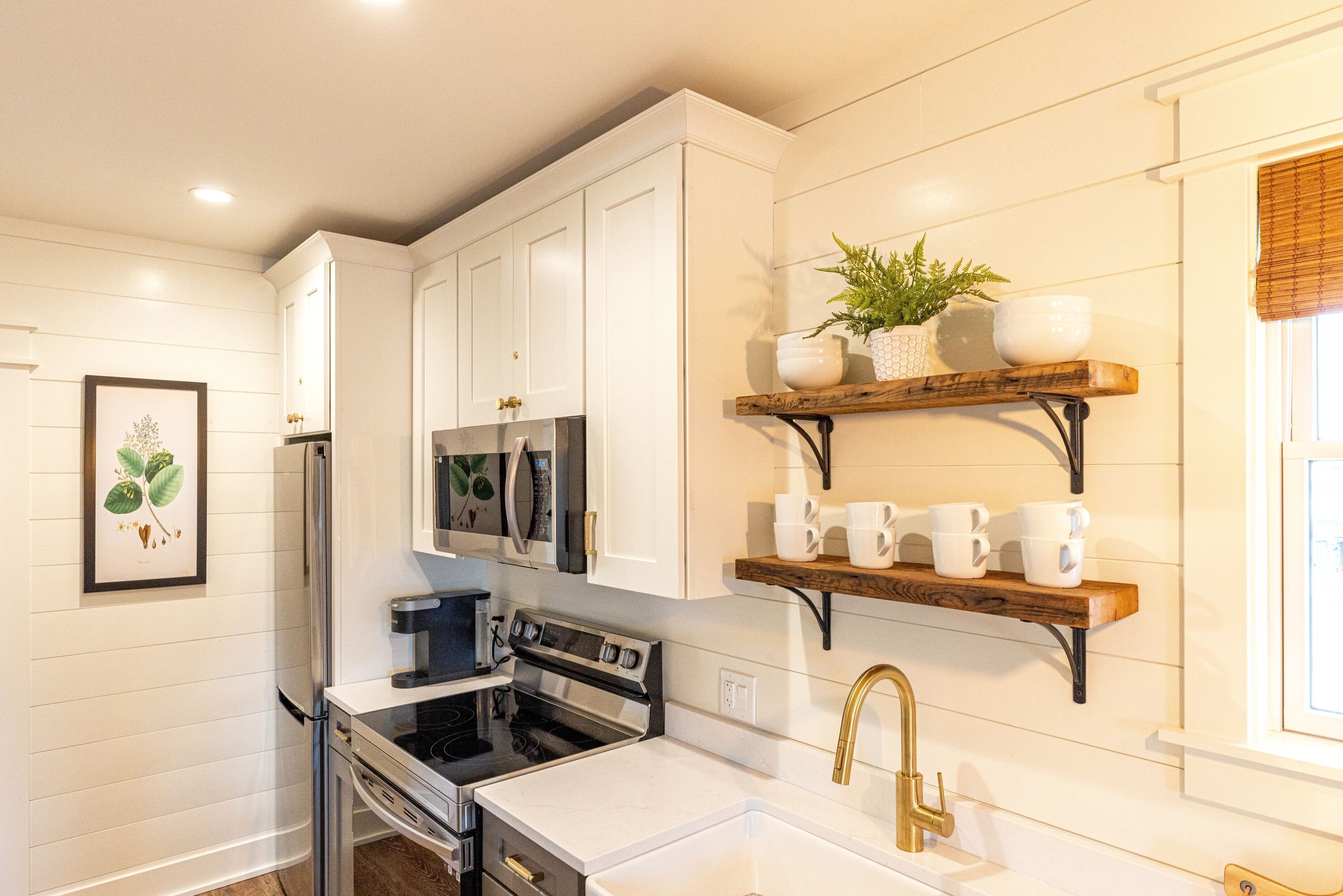 Una cocina con muebles blancos, un refrigerador de acero inoxidable, una estufa negra y un estante de madera con tazas y una planta.