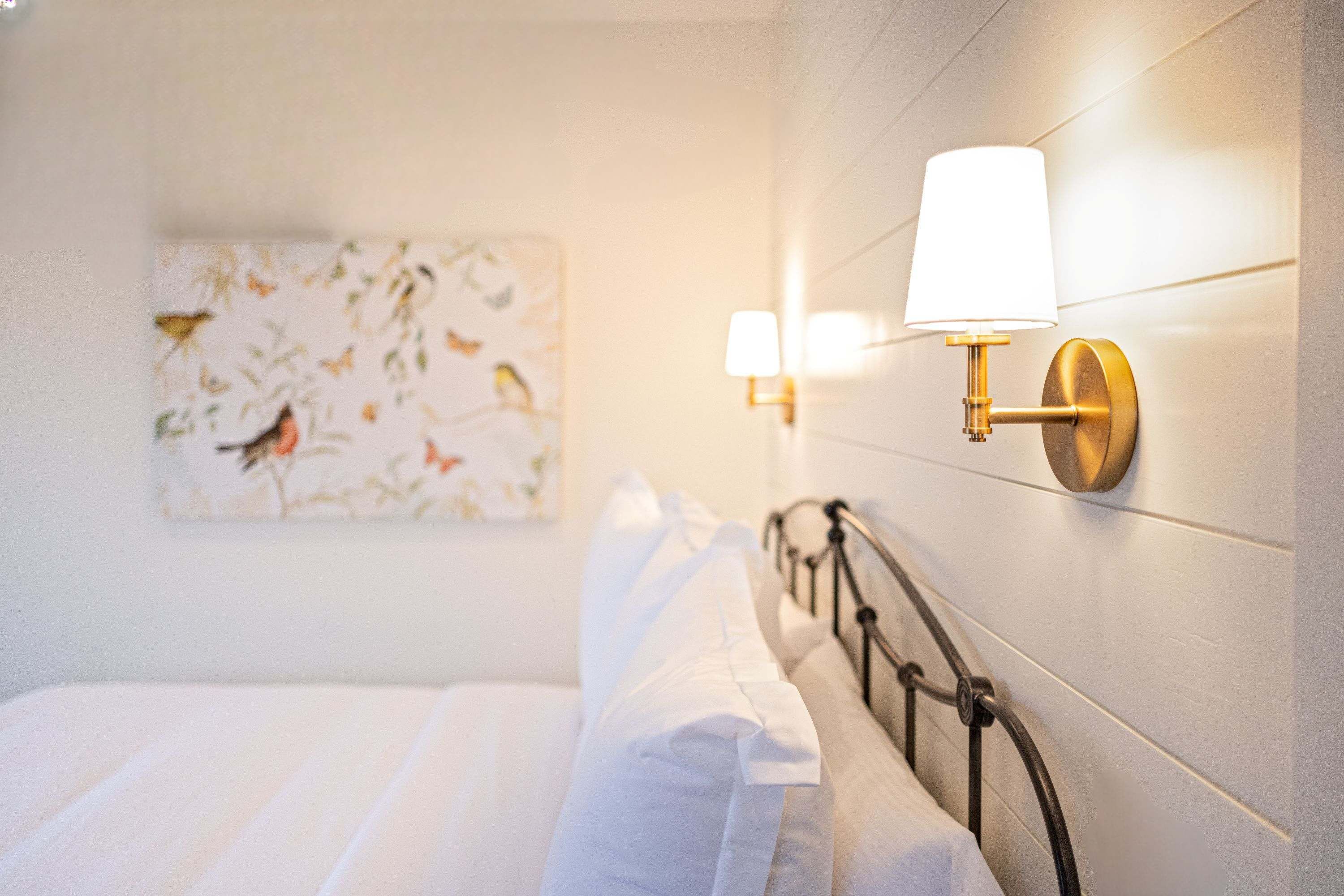 A neatly made bed with white linens, a wall-mounted light fixture, and a floral artwork on the wall.