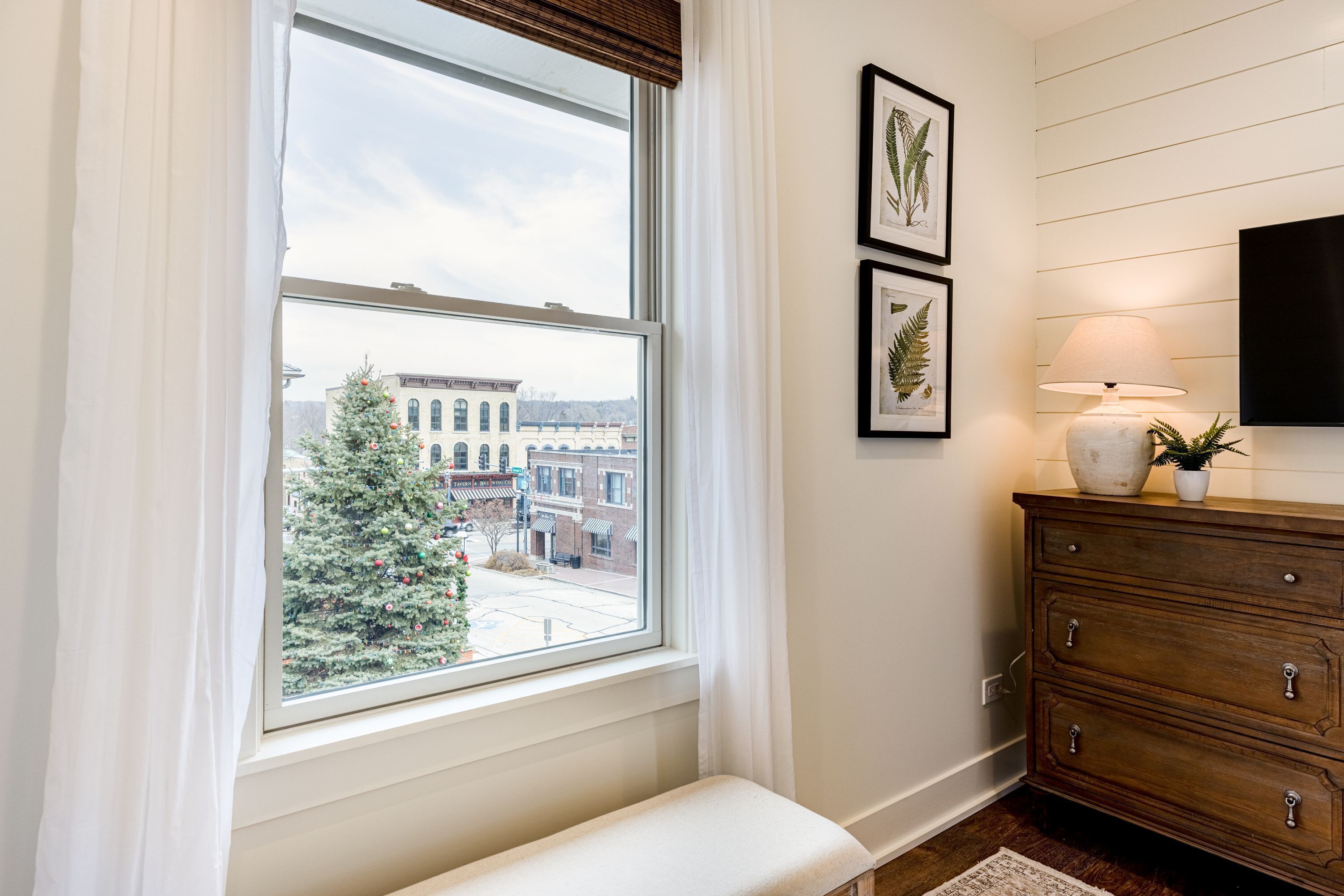 A room with a large window, a bench, a wooden dresser, and framed botanical prints on the wall.