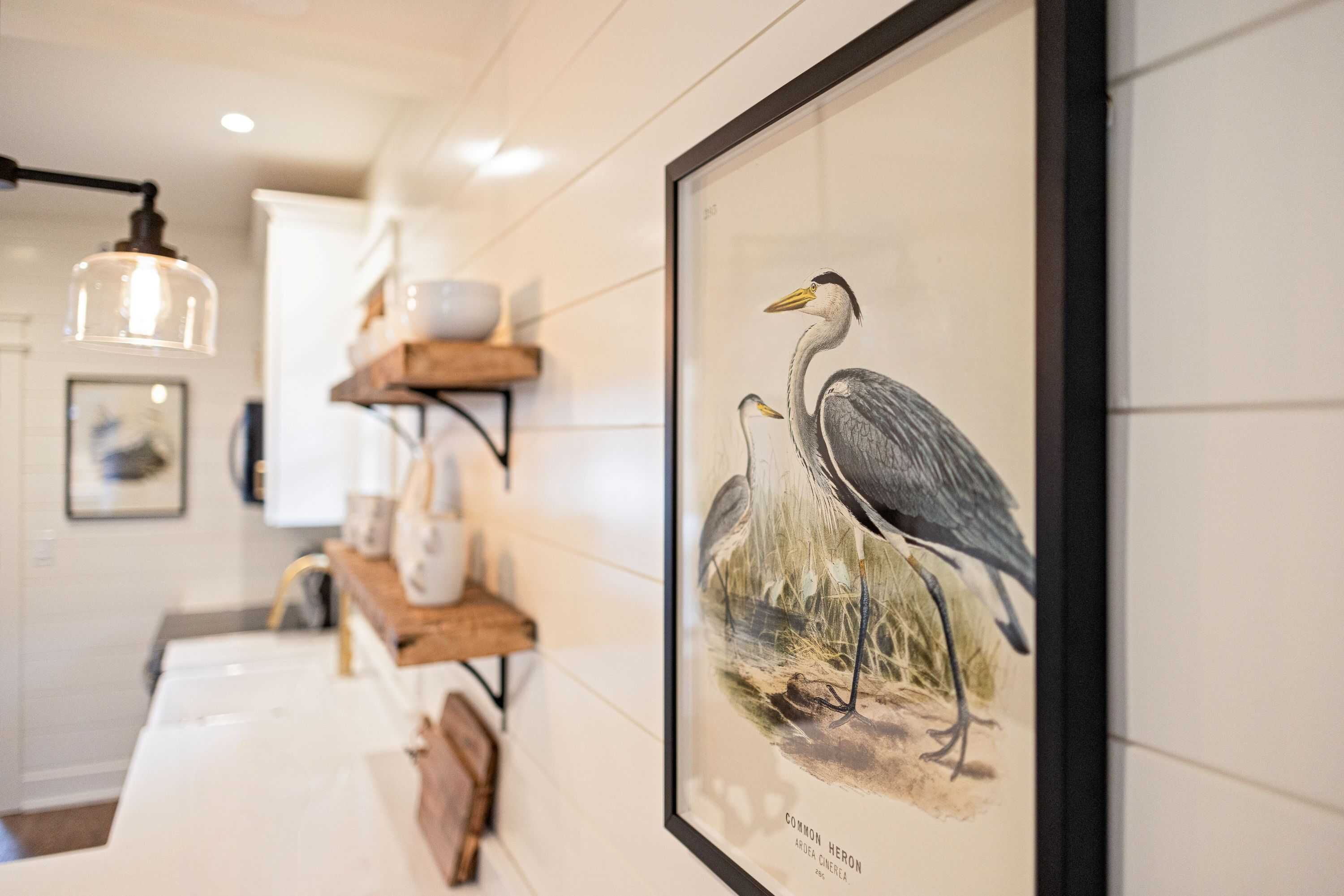 Una ilustración de pájaro en un marco en la pared, una lámpara colgante y un estante de madera con objetos.