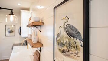 Una ilustración de pájaro en un marco en la pared, una lámpara colgante y un estante de madera con objetos.